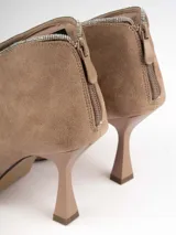 Botín color beige de gamuza con punta fina y taco de aguja. El borde superior está adornado con strass plateados.