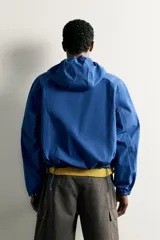 Cazadora técnica relaxed fit de color azul, con cuello subido, capucha ajustable, bolsillos frontales de plastrón con solapa, cierre de cremallera oculta y puños elásticos.