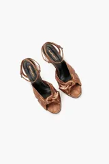 Sandalia de noche color bronce metalizado con taco alto y plataforma. Presenta una capellada con nudo fruncido y tira fina ajustable en el tobillo con hebilla.