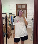 Remera blanca de manga corta y corte holgado, con un estampado frontal en tonos rosados que incluye una palmera y texto.