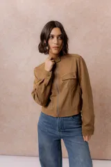 Chaqueta de gamuza sintética color tostado, con cuello mao, cierre frontal con cremallera y bolsillos frontales con solapa. Presenta detalles de broches metálicos y puños ajustables.