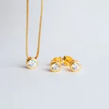 Set de joyería compuesto por un collar con dije de punto de luz y un par de aros tipo caravanas a juego. Ambas piezas presentan circonias engarzadas en bases circulares con baño de oro.
