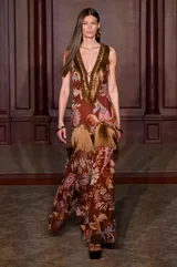 Vestido largo marrón de estilo boho con estampado paisley y floral en tonos tierra, rosado y celeste. Presenta escote en V profundo decorado con flecos dorados, pedrería y cordones entrecruzados, además de detalles de flecos en la zona de la cadera.