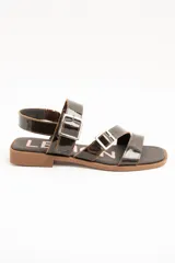 Sandalias de cuero charolado negro con dos tiras con hebillas metálicas plateadas y tira en el talón. Suela plana color marrón.