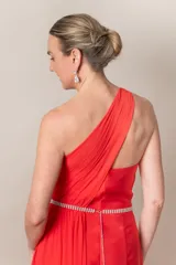Vestido largo de fiesta color rojo, de corte recto y un hombro descubierto. Presenta una pieza de gasa que nace del hombro, formando el bretel y cayendo a lo largo del vestido.