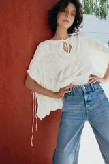 Top blanco de cuello redondo y mangas tipo capa, con textura fruncida, bajo asimétrico, lazos decorativos en el escote y en los laterales, y cierre posterior con botón.