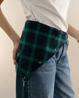 Bandana triangular de tela con estampado a cuadros en tonos verde y azul marino. Presenta ojales metálicos distribuidos en los bordes para la personalización con accesorios y un aro metálico en la punta inferior.