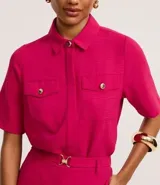 Camisa color fucsia de tejido airflow, con cuello camisero, mangas cortas con pliegues, cierre frontal con botones metalizados y bolsillos con solapa en el pecho.