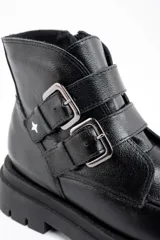 Bota corta de cuero negro con suela track dentada y dos correas decorativas con hebillas metálicas en el lateral. Presenta un diseño de puntera cuadrada y cierre lateral.