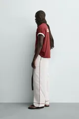 Camiseta blanca de corte relajado (relaxed fit) con cuello redondo y manga corta. Presenta un bolsillo plastrón en el pecho y un estampado de texto en color marrón rojizo en la espalda que dice "SOMETIMES SÍ, SOMETIMES BYE" (reemplazando la 'O' de 'SOMETIMES' por un corazón).