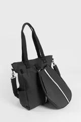 Bolso tipo tote negro confeccionado en PVC y poliéster, con compartimento principal con cierre, bolsillos laterales y bolsillo trasero con cierre. Incluye asas superiores de mano, correa ajustable removible y un estuche removible para paleta de pádel con detalle de franja vertical blanca.