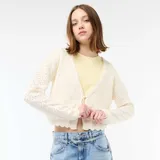 Chaqueta de punto con textura de croché en color beige, de corte corto y mangas largas. Presenta un cierre frontal mediante una lazada fina y bordes con acabado festoneado.
