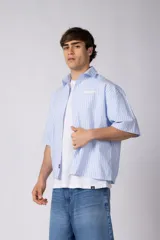 Camisa de manga corta a rayas beige y blancas, de corte boxy y liviana, con cuello camisero, cierre frontal de botones y pequeño texto bordado "BUILD IT BOLD" en el pecho.