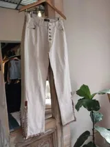 Pantalón de jean blanco con corte acampanado, cintura alta, detalles trenzados y aplicaciones de cuero en la parte inferior.