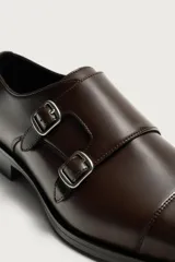 Zapato de vestir tipo monk de color negro, con detalle de doble hebilla metálica plateada en el empeine, puntera redondeada y suela con ligero tacón.