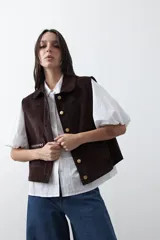 Chaleco de pana color marrón oscuro, con cuello camisero, cierre frontal de botones metálicos dorados y dos bolsillos frontales con detalle de bordado geométrico.