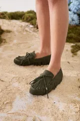 Mocasines de gamuza color tostado, con diseño clásico, flecos alrededor del calzado y en la capellada, y cordón decorativo con detalles metálicos dorados.