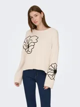 Sweater de punto color crema con cuello redondo y mangas largas. Presenta un diseño con bordados florales en color negro ubicados en el pecho y en una de las mangas.