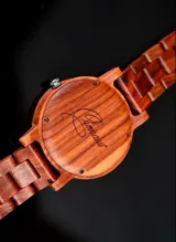 Reloj de pulsera con caja, esfera y malla de madera de sándalo rojo, con marcadores y manecillas negras. Presenta un diseño circular con corona lateral.