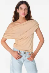 Blusa asimétrica color beige de poliéster con spandex, con diseño drapeado envolvente que deja un hombro al descubierto y manga corta caída del otro lado.