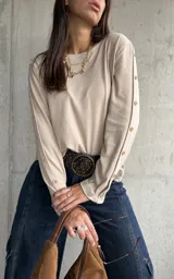 Remera de lanilla color beige, de corte holgado y manga larga. Presenta una hilera de botones decorativos a lo largo de toda la parte externa de las mangas.
