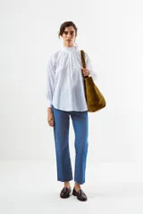 Blusa de algodón blanco con diseño de rayas verticales y textura de pequeños puntos en relieve. Presenta cuello alto con volados, cierre frontal con botones forrados y mangas largas abullonadas con puños ajustados.