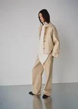 Chaqueta corta de algodón color beige, de corte holgado, con cuello clásico, cierre frontal de botones y bolsillos ribeteados. Cuenta con mangas largas amplias con puños ajustables mediante tiras y botones.