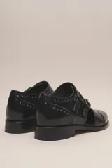 Zapato tipo monk strap con aberturas laterales, confeccionado en cuero vacuno metalizado color bronce. Presenta detalles de perforaciones estilo brogue en la puntera y el talón, y doble hebilla metálica en el empeine.