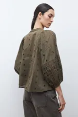 Blusa color verde oliva de manga 3/4 con volumen, cuello mao con abertura en V y botones. Presenta bordados y detalles de broderie calado en el canesú frontal y en las mangas. El cuerpo tiene un corte peplum con frunces.