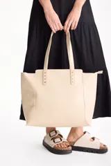 Cartera tipo tote bag color beige con diseño texturado efecto croco, asas dobles y detalles de tachas metálicas en la parte superior.