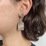 Par de aros colgantes con base negra, cadena plateada y dije transparente con dibujo de ojo rosa.