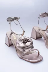 Sandalias de cuero color cobre con taco cuadrado y tira fina sobre los dedos. Presentan una flor grande en el empeine y tiras finas para atar al tobillo.