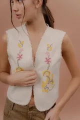 Chaleco color crudo sin mangas, con escote en V y bordados artesanales florales en tono amarillo, rosa y marrón en el frente. Presenta detalles de pasamanería con pompones pequeños en los bordes.