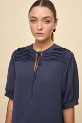 Blusa de manga corta color negro, con calce relajado, escote redondo con abertura y lazo para anudar. Presenta detalles de tablas horizontales en la parte superior que aportan textura al diseño.