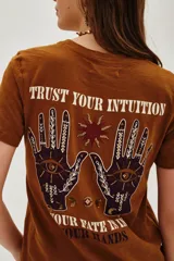Remera de algodón color marrón con estampa gráfica en la espalda que incluye manos, un ojo y la frase 'Trust your intuition, your fate is in your hands'.