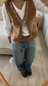 Chaleco de gamuza color camel con bordados decorativos en color blanco y cierre frontal con cordones y apliques metálicos.