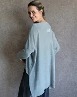 Buzo de tejido de punto en color gris claro, con cuello redondo, mangas largas con hombros caídos y diseño de corte asimétrico con la parte trasera más larga que la delantera.