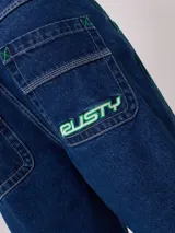 Bermuda de jean de mezclilla negra con efecto lavado, corte recto y costuras en contraste de hilo blanco. Presenta un parche bordado con el logo "Rusty" en color celeste en el bolsillo trasero derecho.