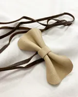 Gargantilla tipo choker con dije en forma de moño de cuero color beige y tiento de cuero marrón para atar. El dije mide 8,5 x 4,5cm y el tiento mide 94cm de largo.