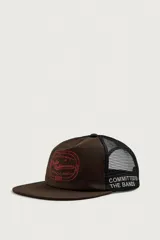 Gorra trucker marrón con visera plana y red negra en la parte trasera. Cuenta con un bordado rojo en el frente con la inscripción "Indie Behaviour 1975" y otro bordado blanco en el lateral que dice "Committed to the bands".
