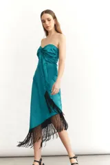 Vestido midi sin mangas color turquesa, con escote strapless y detalle de nudo frontal. El ruedo presenta un corte asimétrico con flecos negros largos.