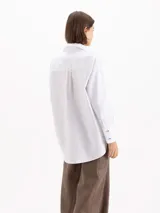 Camisa holgada de poplin blanco, con cuello clásico, mangas largas con puños anchos y cierre frontal con botones. Presenta detalles de apliques metálicos en las puntas del cuello y sutil bordado en el delantero.