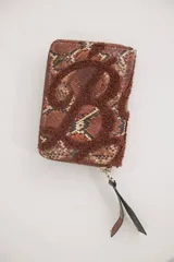 Billetera rectangular con cierre perimetral, confeccionada en material con estampado de piel de serpiente en tonos marrones y naranjas. Presenta una letra B grande aplicada en relieve con textura de felpa marrón y un tirador de cierre largo en cuero.
