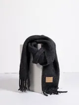 Bufanda de tejido grueso en color gris oscuro, con flecos en los extremos y etiqueta de cuero sintético con el logo de la marca en una de las puntas.