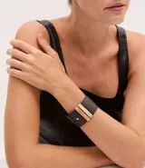 Pulsera ancha de material sintético color marrón oscuro, con diseño de cinta larga y dos tiras metálicas doradas aplicadas en el centro. Cuenta con un cierre de pino ajustable.