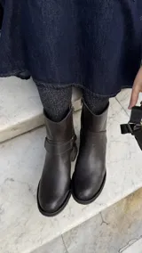 Botas cortas de cuero marrón, con taco cuadrado y caña baja. Presentan una tira con hebilla metálica decorativa alrededor del tobillo y cierre lateral.