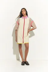 Vestido corto tipo túnica color crema con bloques de color beige en hombros y laterales. Presenta cuello camisero rosa, cierre frontal con línea roja y terminación con franja amarilla. Detalles de líneas turquesas y fucsias en hombros.