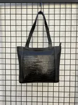 Cartera tipo tote de cuero negro, con doble asa larga para hombro y bolsillo frontal con textura efecto cocodrilo y marca grabada. Presenta costuras visibles en contraste.