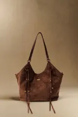 Bolso tipo shopper de gamuza vegana color marrón, con tiras colgantes decorativas a los lados y placa metálica frontal con el logo de la marca. Cuenta con asas largas de hombro.