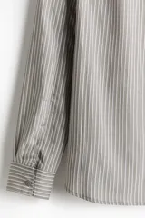 Camisa de manga larga con cuello inglés y corte regular, confeccionada en tejido suave con rayas verticales grises y blancas. Presenta tapeta lisa, canesú con pliegue de caja en la espalda, puños abotonados y bajo levemente redondeado.